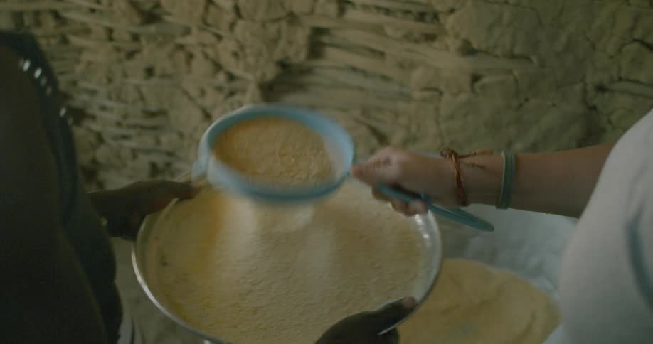 Slow-motion close-up of two people sifting fine yellow grain through a sifter onto a large silver platter