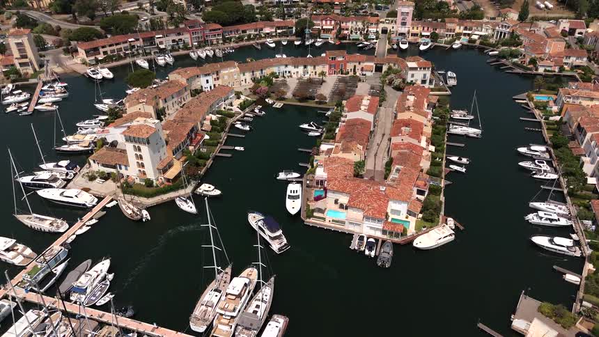 An aerial drone slowly flies over a picturesque European-style marina filled with docked yachts and surrounded by colorful waterfront homes