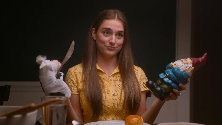 A young woman at a dinner table stages a bizarre and violent fight between a cow hand puppet and a garden gnome