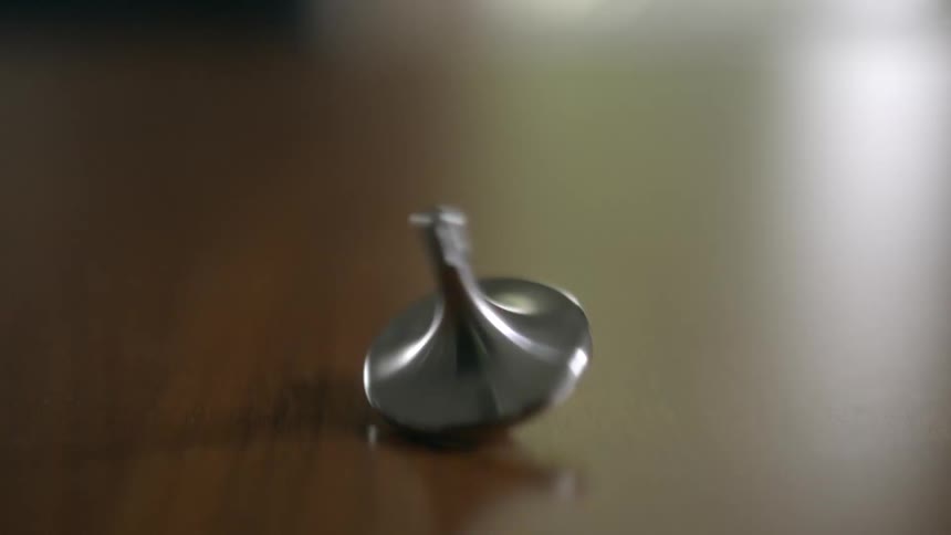 A metal spinning top totem spins on a wooden table, eventually losing momentum and wobbling