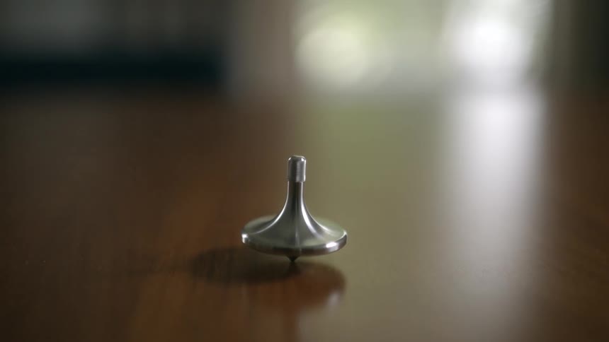 A person's hand spins a silver metal totem top on a reflective wooden table