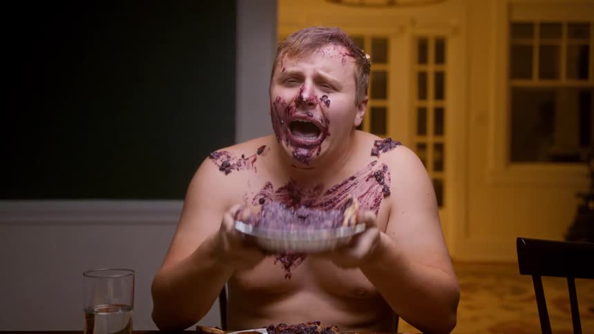 A shirtless man cries hysterically while smashing a blueberry pie all over himself at a dining table