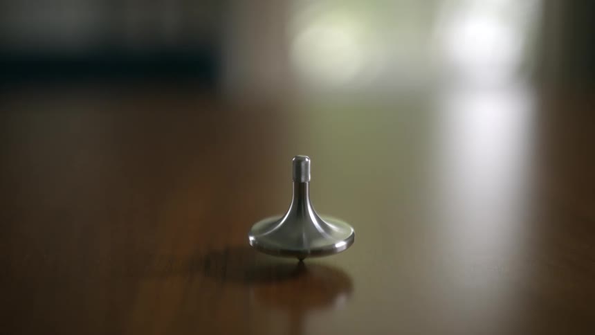 A sleek, metallic spinning top rotates endlessly on a smooth, dark wooden table in a mesmerizing close-up shot