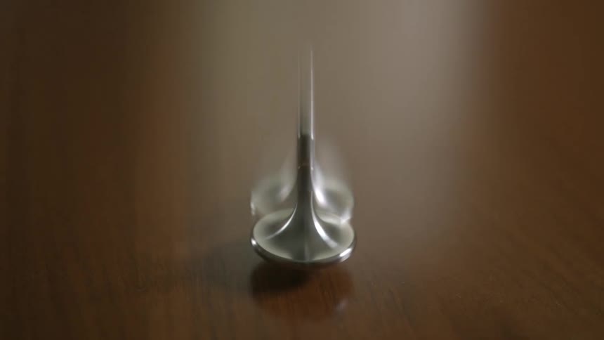 A sleek silver spinning top rotates and wobbles in a seamless loop on a dark wooden surface