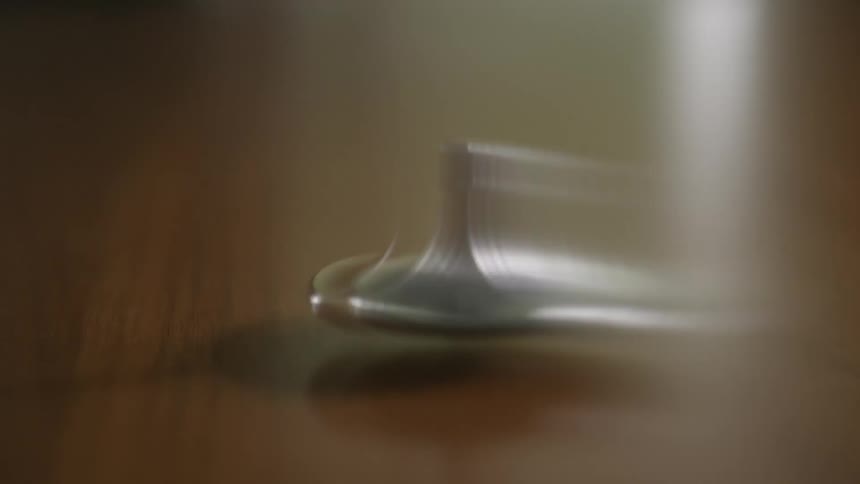 A close-up shot of a small, silver spinning top continuously rotating on a smooth, reflective wooden surface