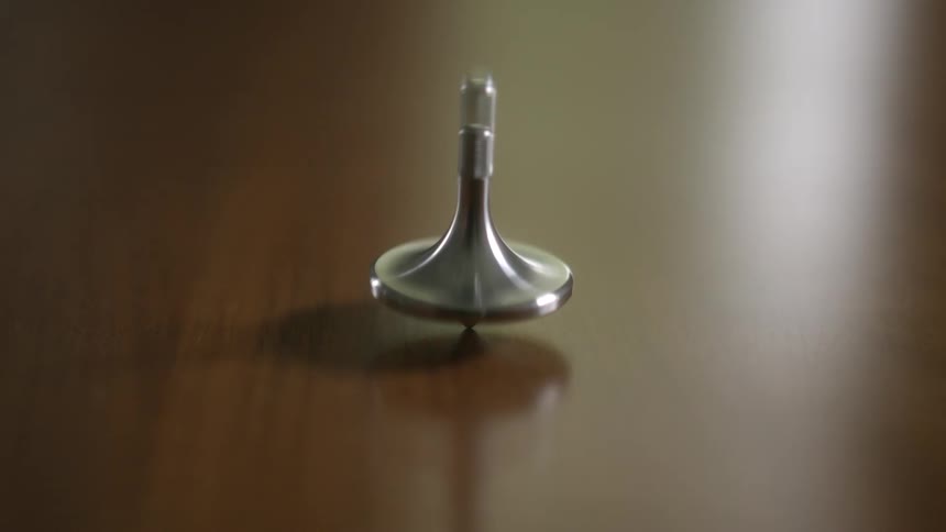 A metal spinning top spins and wobbles on a smooth wooden surface in a mesmerizing close-up