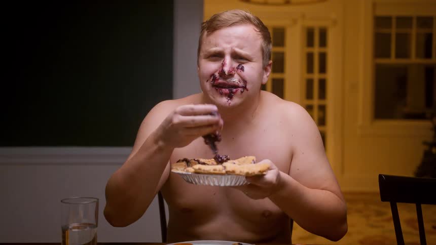 A shirtless man ravenously eats a whole blueberry pie with his hands, making a complete mess of his face