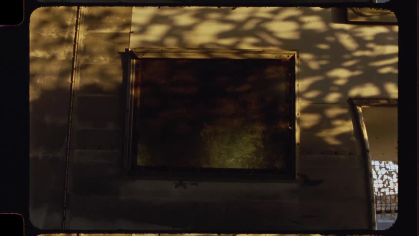 Dappled sunlight and tree shadows play across the dirty window of an old metal trailer in a vintage-style shot