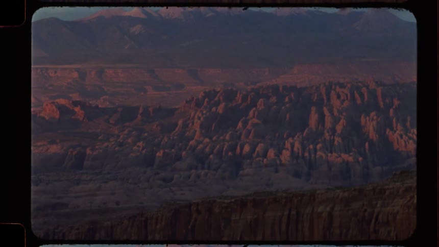 A vintage film shot tilts up to reveal the stunning, sun-kissed rock formations of a vast desert canyon with mountains in the distance