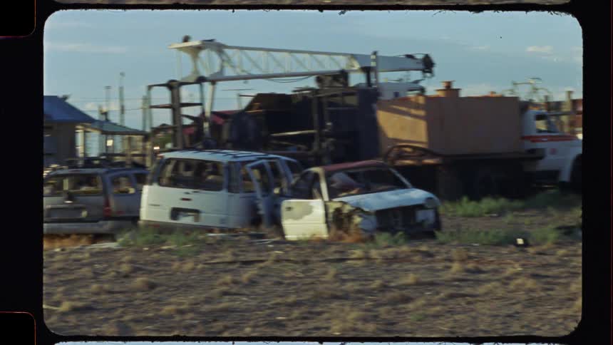 Archival film footage pans across a desolate junkyard with abandoned and wrecked cars