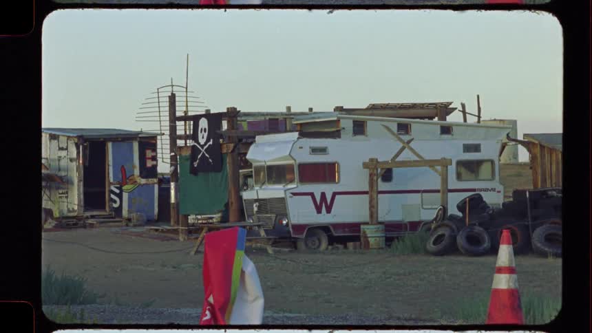 A pirate flag hangs over a weathered RV and makeshift sheds in a desolate, off-grid encampment