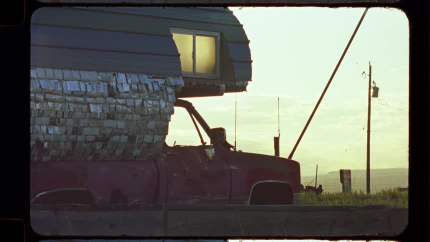 A static vintage film shot of a beat-up red pickup truck with a camper shell parked in a desolate, rural landscape at dusk