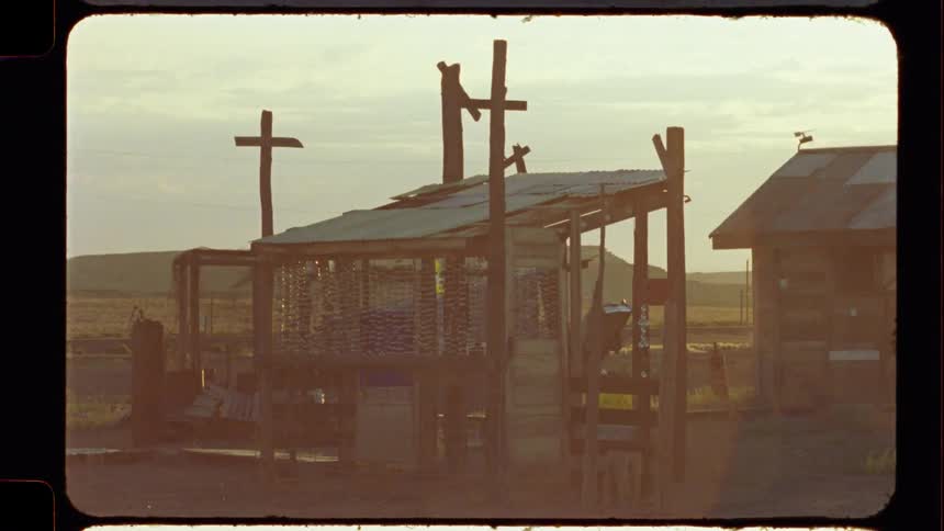 A rustic wooden structure and a cross stand in a desolate, rural landscape during a hazy sunset, captured on vintage film