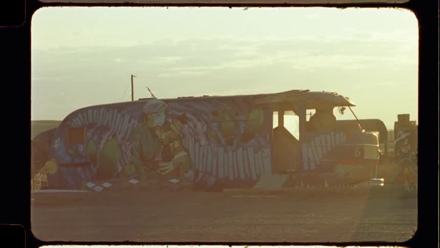 An old, abandoned bus covered in colorful graffiti sits motionless in a dusty, barren landscape during a hazy sunset, captured on vintage film