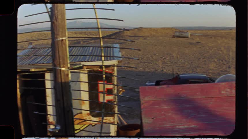 A gritty, sun-flared pan across a desolate desert junkyard at sunset, captured on vintage film