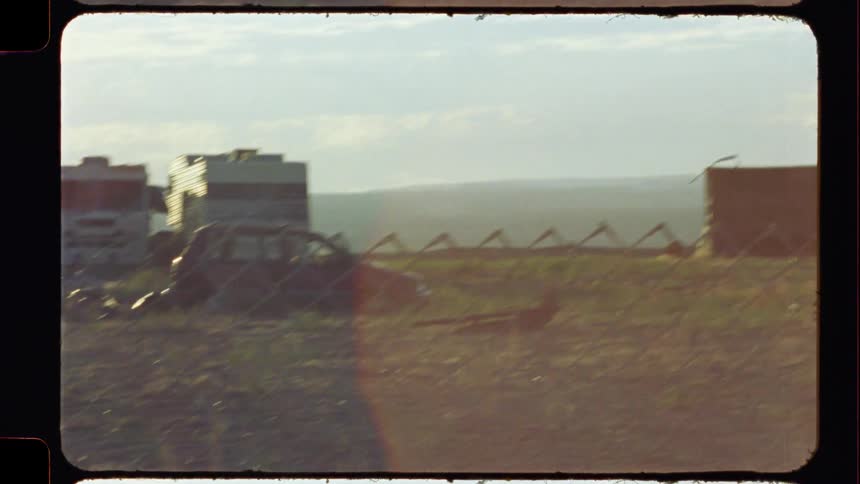 Driving past a desolate junkyard and a small, impoverished village with abandoned cars, captured on vintage 8mm film