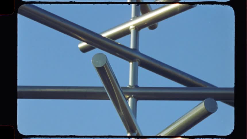 A vintage 8mm film captures an upward tilt of a modern metallic sculpture against a clear blue sky