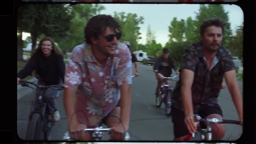 Vintage film footage captures a group of smiling friends enjoying a leisurely bike ride through a suburban neighborhood