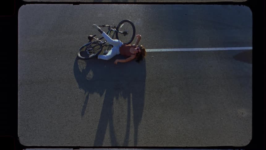 An overhead aerial view shows a young woman lying on the ground after a serious bicycle accident on a paved road