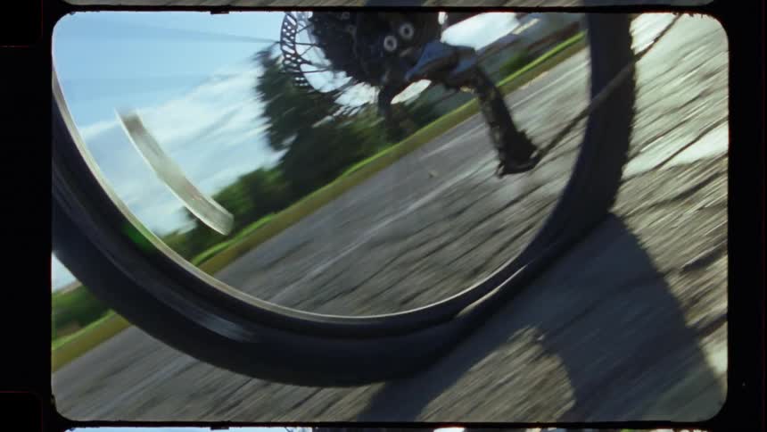 A dynamic close-up of a bicycle wheel spinning down a paved road on a sunny day, captured on vintage 8mm film