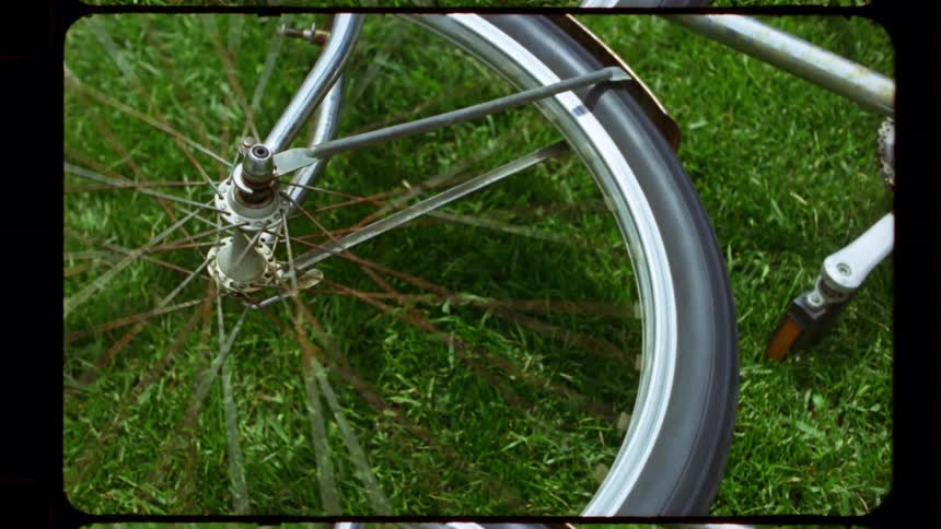 A vintage film effect shows a close-up of a rusty bicycle wheel spinning on the grass