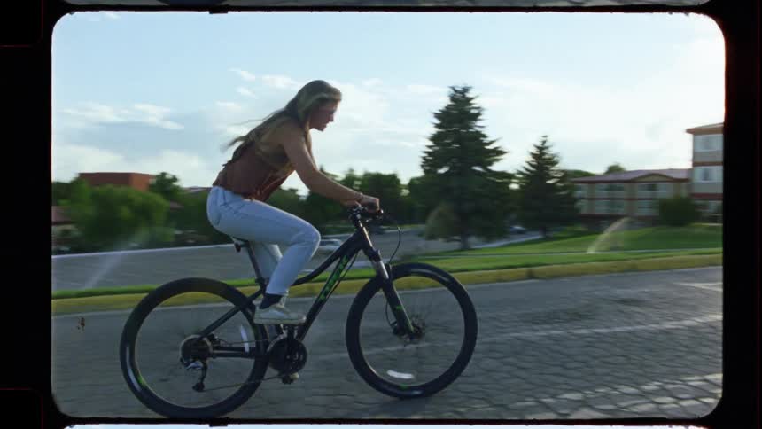 A young woman rides her bicycle in slow motion down a cracked street at golden hour, with a distinct film look