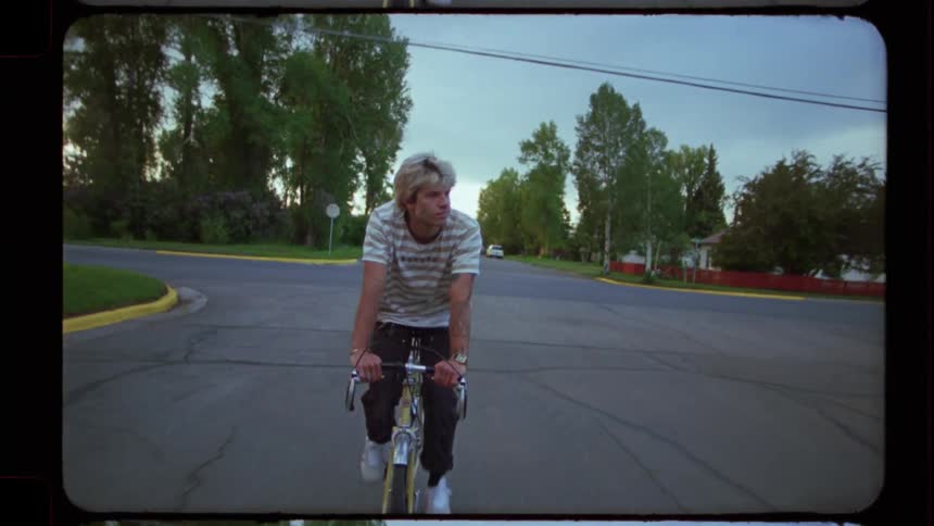 Vintage film-style footage captures a young man riding his bicycle down a quiet suburban street at dusk