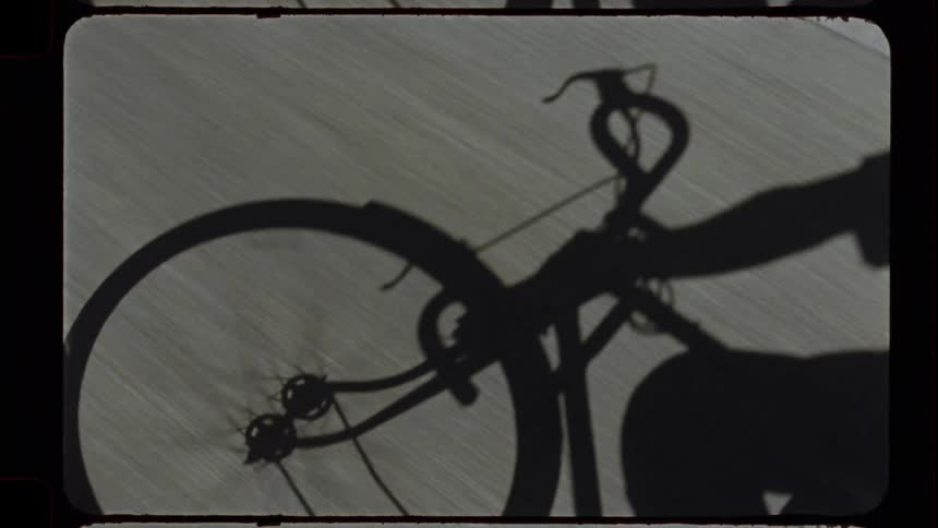 An artistic point-of-view shot from a bicycle shows the cyclist's shadow on the moving pavement in vintage-style film