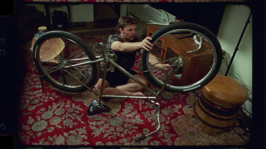 A man barefoot on a red patterned rug works on his upside-down bicycle inside his apartment