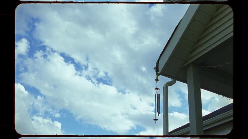 A vintage film-style time-lapse shows fluffy white clouds moving across a blue sky above a house with a wind chime