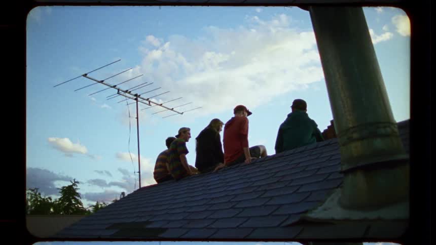 A group of young men sit on a shingled rooftop against a blue sky in this nostalgic, vintage-style footage
