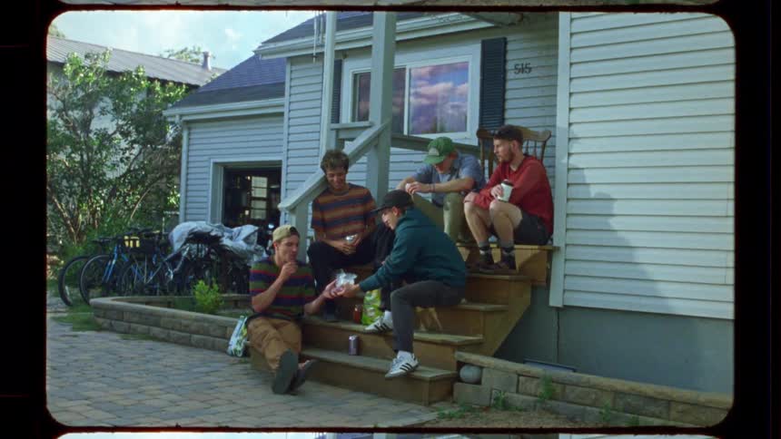 A group of young men in casual streetwear relax and chat on the front steps of a house in this vintage-style film footage