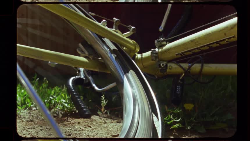 A close-up of a vintage yellow bicycle's wheel spinning on a sunny day in a backyard, captured on retro film