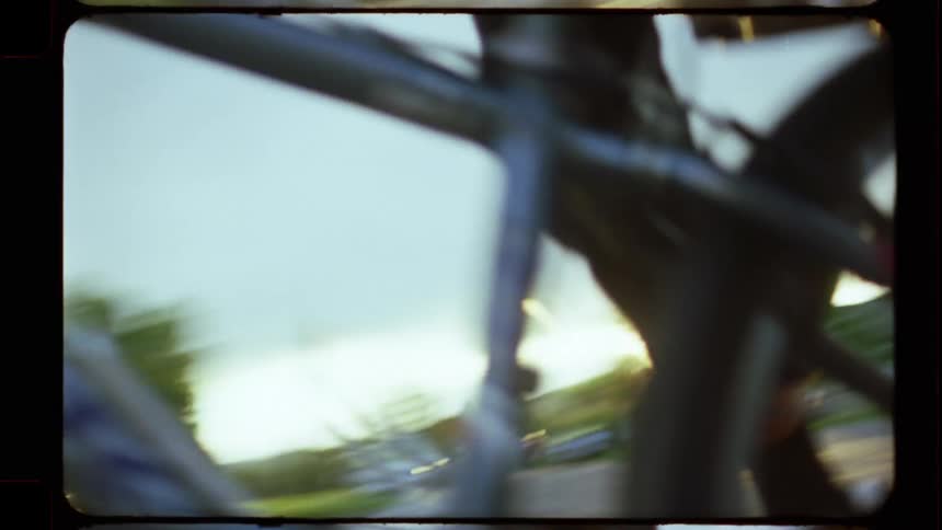 A close-up, shaky, vintage film shot of a young person riding a bicycle through a suburban neighborhood at sunset