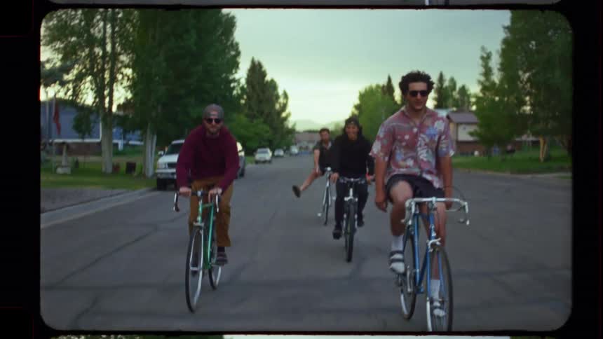 A group of young male friends casually ride their bicycles together down a suburban street in a vintage-style slow-motion shot