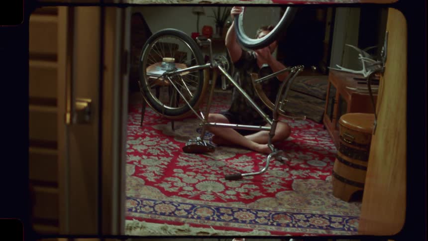 A man with a mustache carefully repairs his bicycle while sitting on a patterned rug in his living room, captured on vintage film