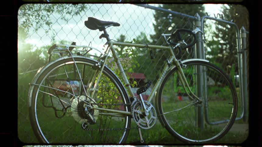 A classic touring bicycle leans against a chain-link fence as the golden hour sun casts a warm, hazy flare in this vintage film-style shot