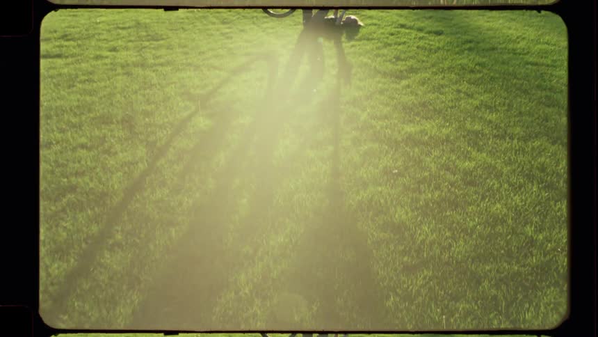 Dreamy film-style footage reveals a person's long shadow on a green lawn before pulling back to show them holding a bike up to the sun