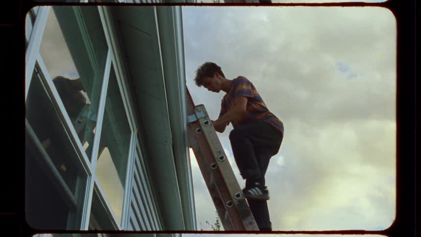 Low angle vintage film shot of a young man climbing a ladder up the side of a house against a cloudy sky