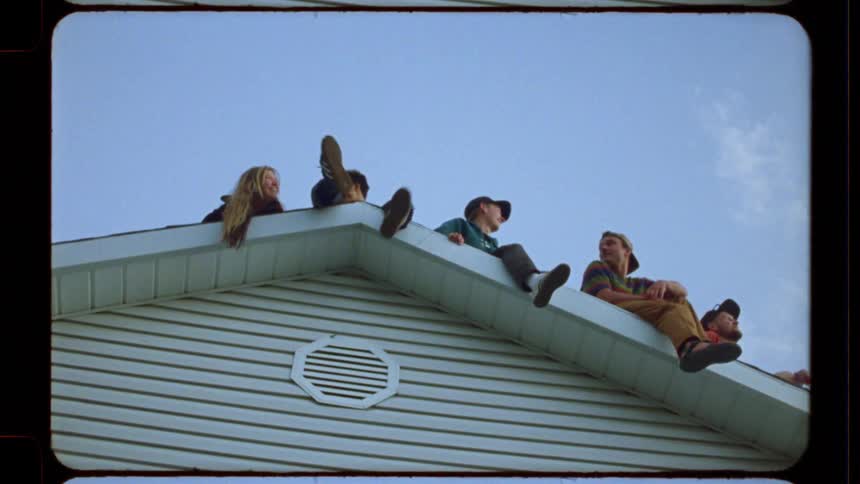 A low-angle, vintage film-style shot of a group of carefree friends hanging out on the roof of a suburban house