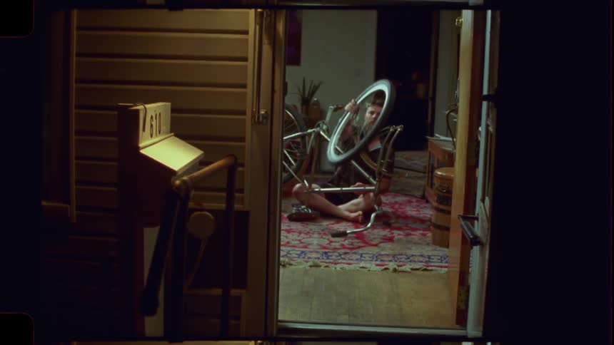 A man seen through an open doorway at night works on fixing his bicycle in his warmly lit apartment