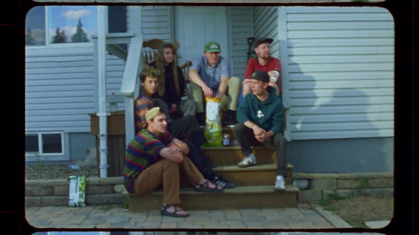 A diverse group of young friends chill on the front porch of a house in this retro-style home movie clip