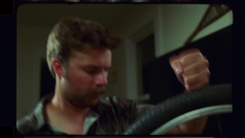 A man with a mustache repairs a bicycle wheel in this retro-style, film-scanned footage