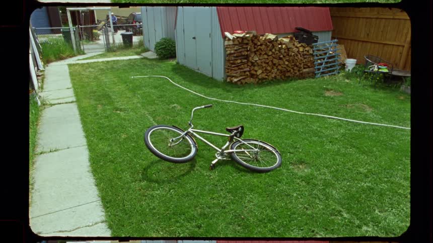 A vintage film-style wide shot of a BMX bike lying on the green grass of a suburban backyard