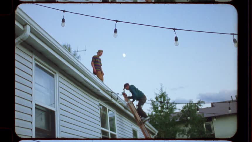 Vintage Super 8 film footage shows two men on a ladder and roof hanging party lights in a backyard at dusk