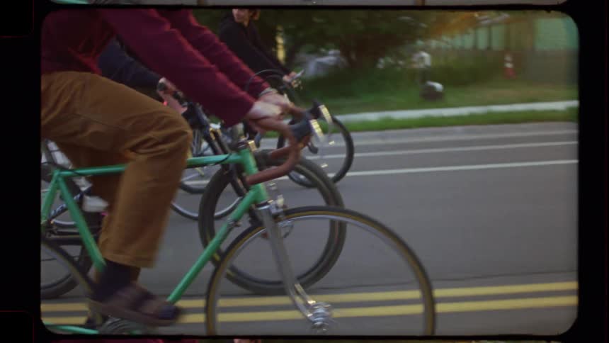 Friends laugh and smile as they ride bikes together through a suburban neighborhood at sunset in this vintage-style footage