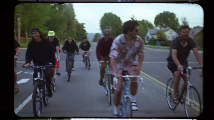 A group of friends smile and laugh while riding their bikes down a neighborhood street in slow motion