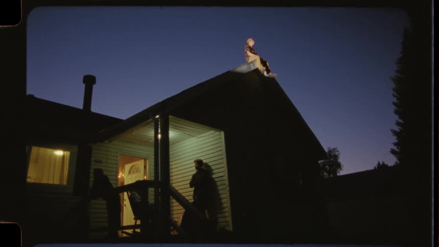 Vintage film style shot of friends hanging out on a suburban porch, with one sitting on the roof as dusk transitions to night