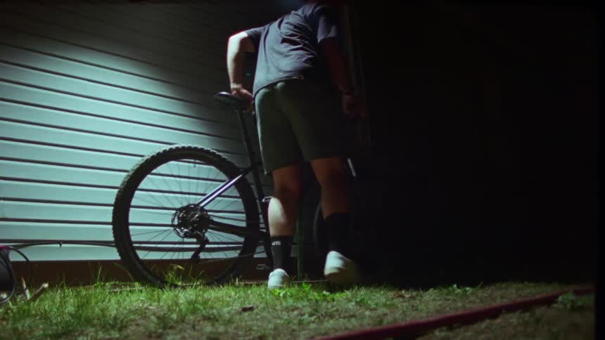 A man walks up to his mountain bike leaning against the side of a house at night and takes it away