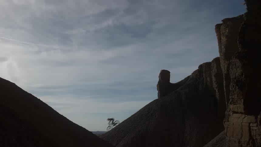 A silhouetted mountain biker performs an impressive jump in slow motion along a steep and narrow desert ridgeline